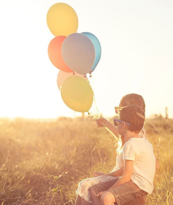 Photograph of two children sitting side by side in the sunset wearing sunglasses and holding a bundle of balloons.  Dravet Syndrome is a seizure disorder that affects children.